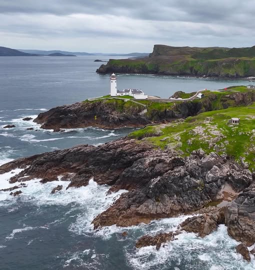 White lighthouse on a rocky coastline with ocean waves and green cliffs under cloudy skies