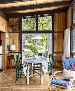 Wood-paneled dining room with farmhouse table, colorful chairs, large windows showing forest view