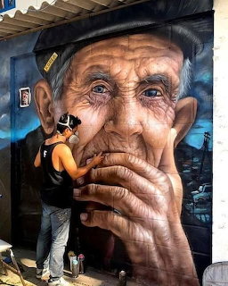 Street artist painting a large realistic mural of an elderly man's face on a wall