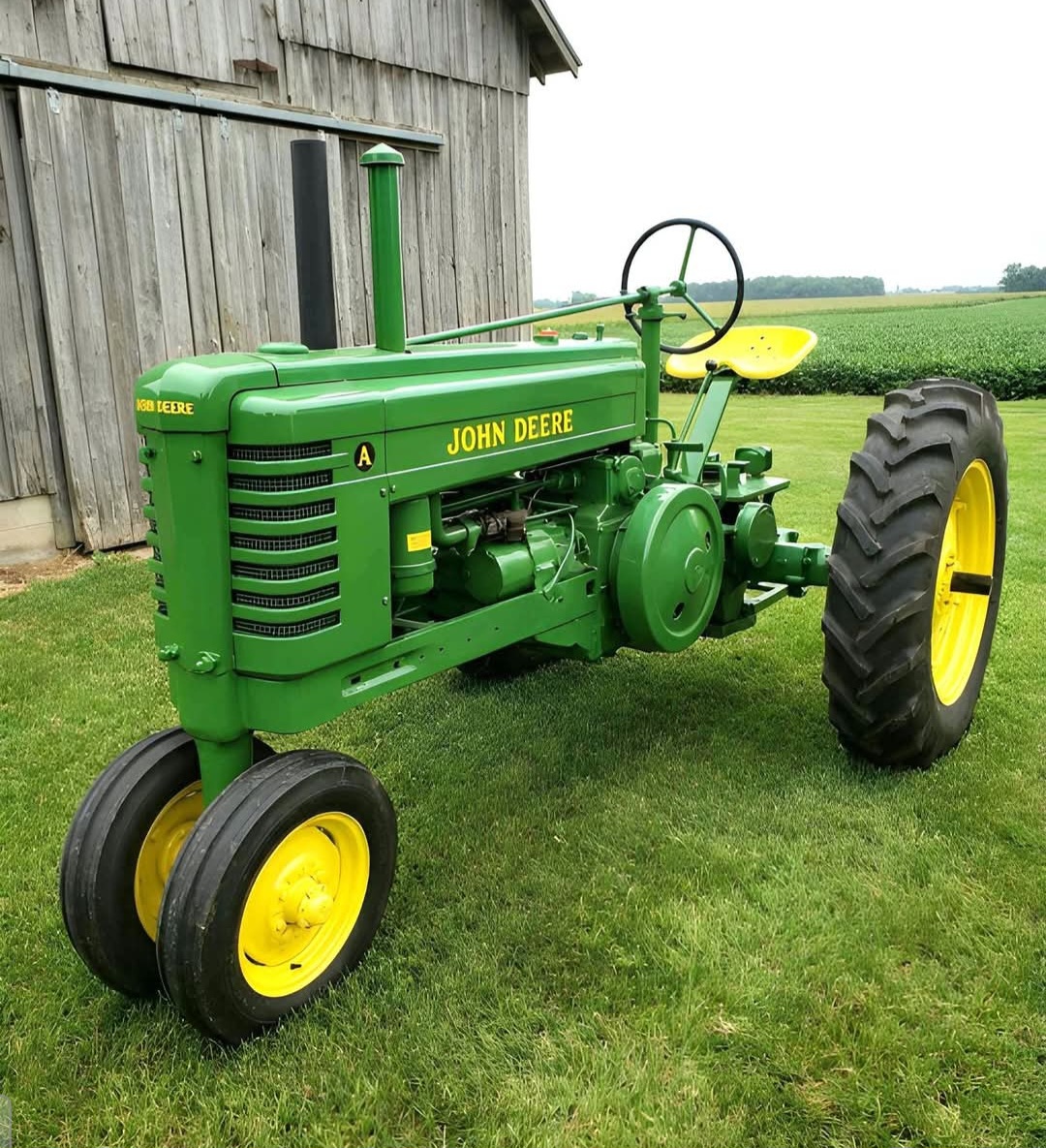 Green John Deere Model A tractor on grass beside a barn and field