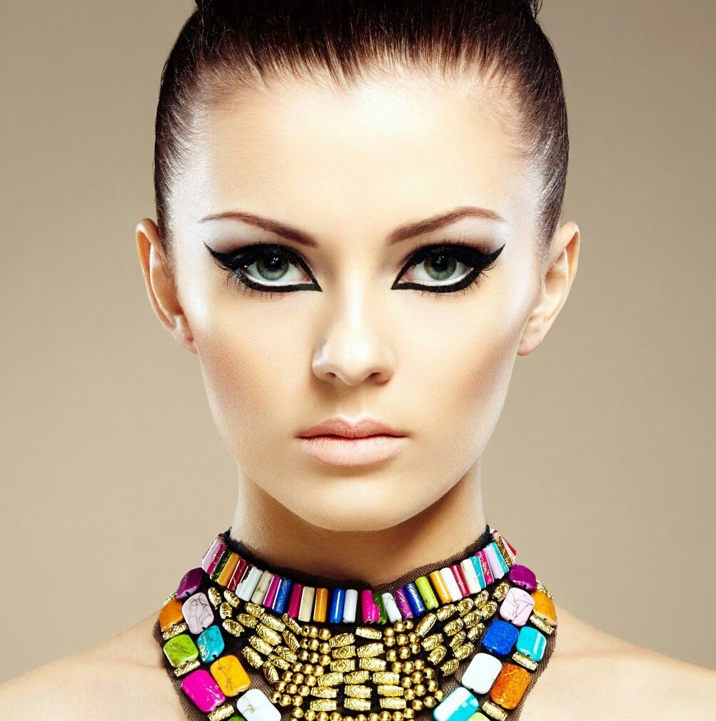 Woman with dramatic black eye makeup and a colorful beaded necklace against a beige background.