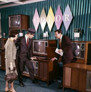 Three formally dressed people in a vintage television showroom with wood-paneled TVs and a COLOR sign