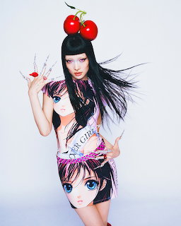 Woman with black hair wearing anime-print outfit and red cherry headpiece posing against white background