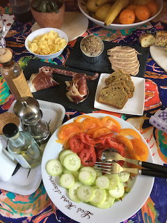 Overhead breakfast table with meats, salmon, cucumber, eggs, bread, and fruit on colorful plates