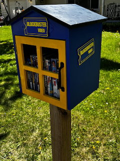 Small blue and yellow Blockbuster Video themed free library box on a wooden post filled with DVDs