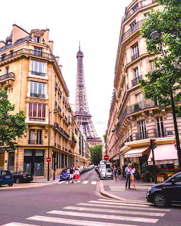 Eiffel Tower framed by cream-colored Parisian apartment buildings along a tree-lined street with pedestrian crossing
