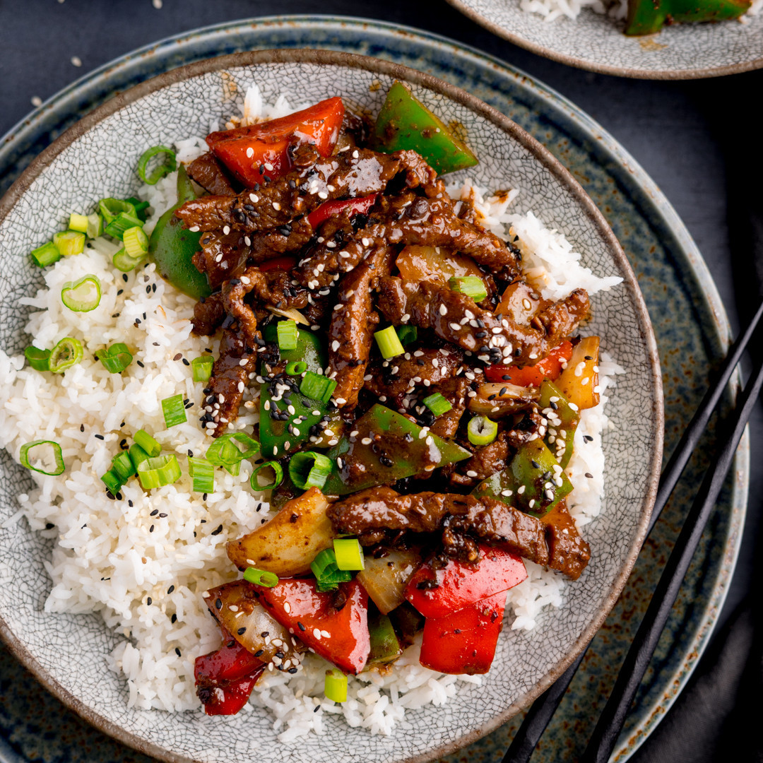 Bowl of white rice topped with sliced beef, red peppers, green onions, and sesame seeds