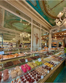 Luxurious bakery interior with ornate turquoise ceiling, chandeliers, and glass display cases full of pastries and desserts