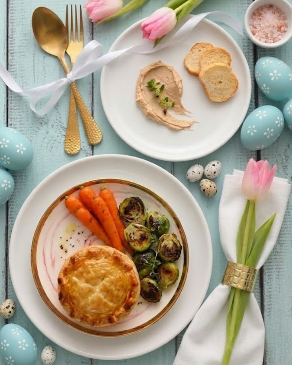 Easter brunch table with roasted carrots, pastries, painted eggs, and pink tulips arranged on light blue background