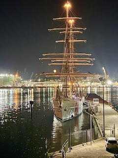 Tall sailing ship with illuminated masts docked at night harbor with city lights in background