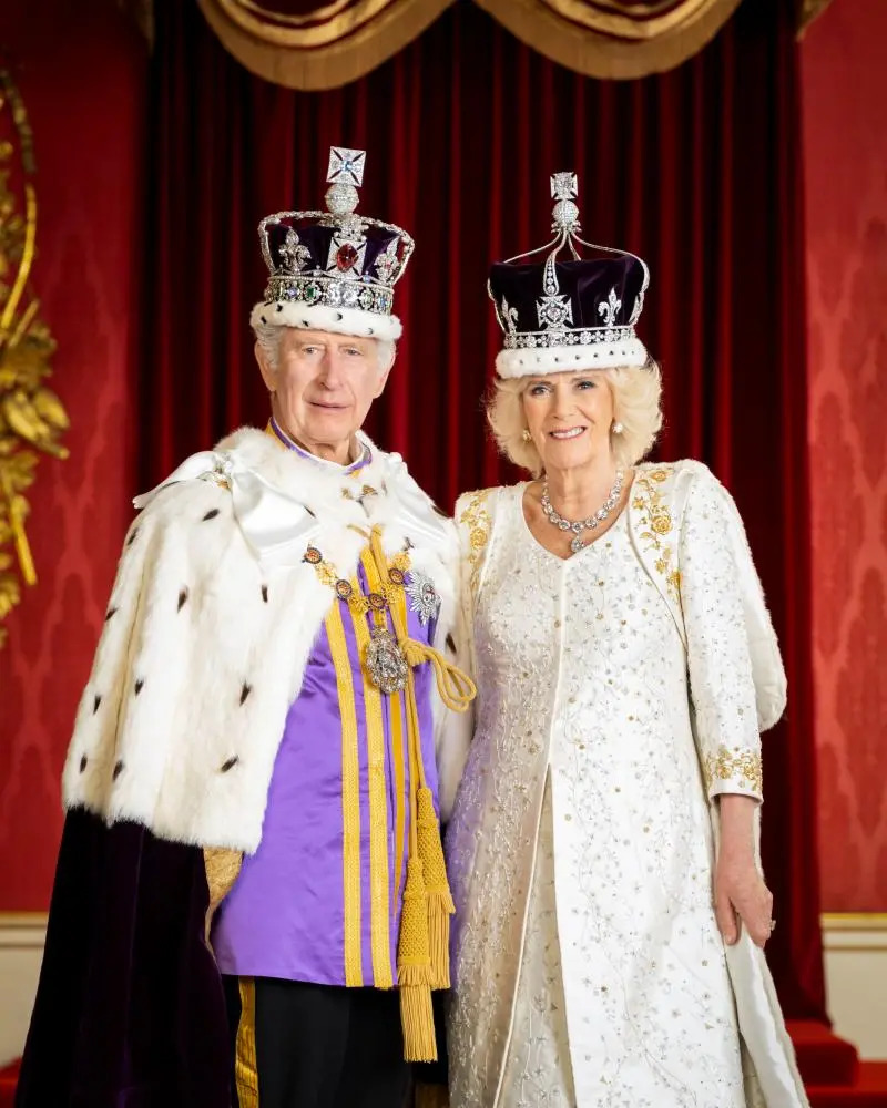 King Charles III and Queen Camilla in coronation robes and crowns standing before red curtains
