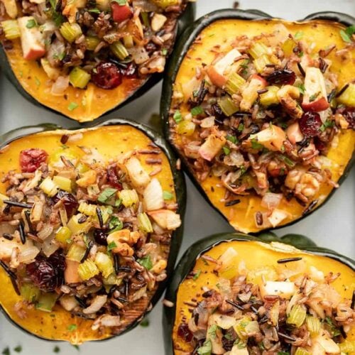 Four roasted acorn squash halves topped with wild rice, cranberries, diced apples, and nuts on a baking sheet