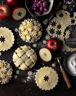 Overhead view of pie crusts, pastry flowers, apples, and baking tools on a dark wooden surface