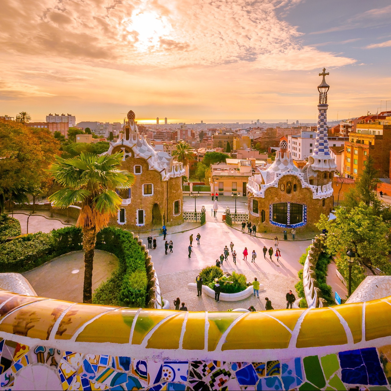Park Güell in Barcelona with mosaic railings, ornate buildings, palm trees, and a sunset sky