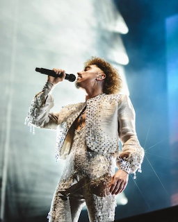 Male rock singer with curly hair in studed silver outfit singing into microphone on stage