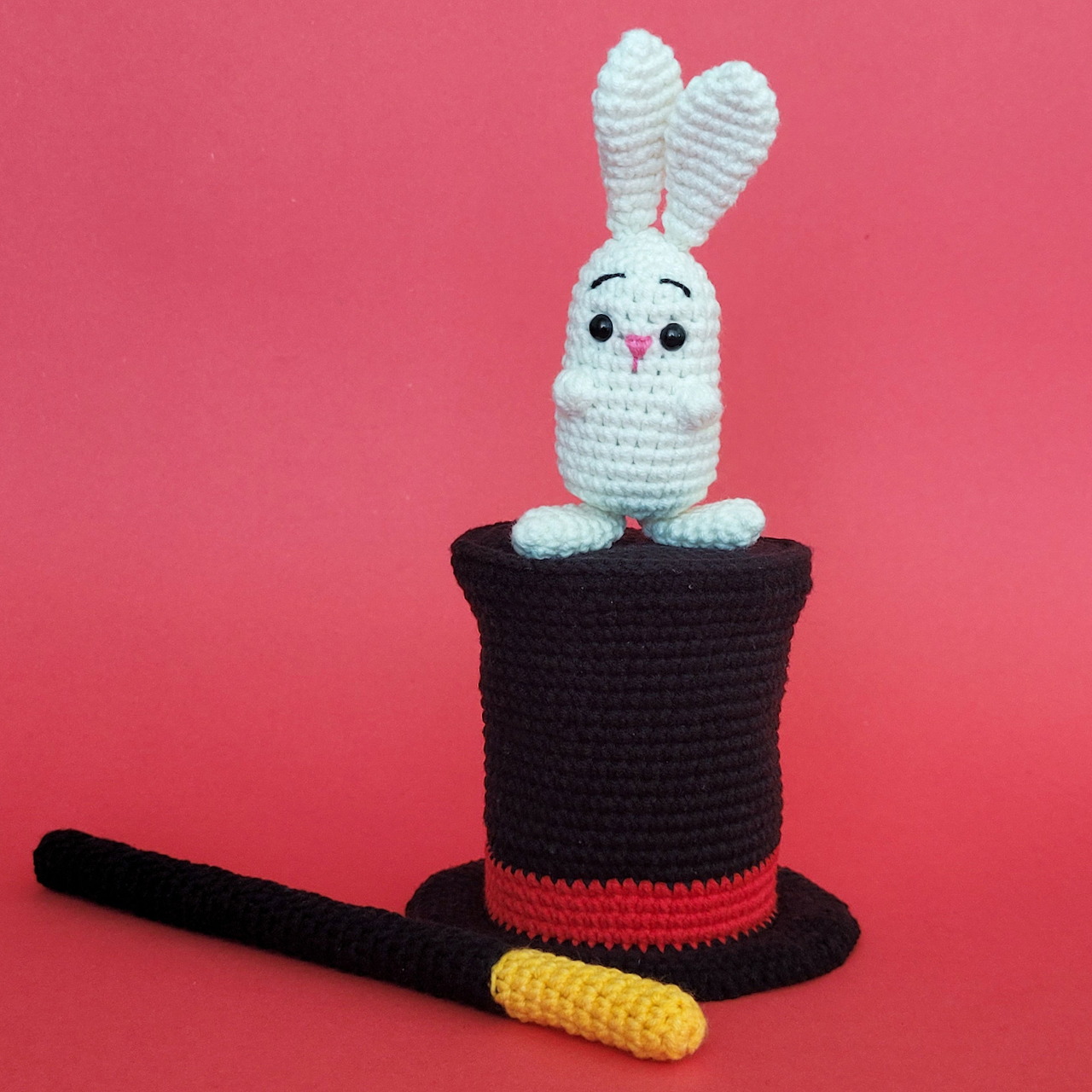 White crocheted bunny in black magician's top hat with wand on pink background