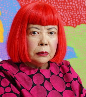 Elderly woman with bright red bob wig wearing purple and black polka dot top in front of colorful art
