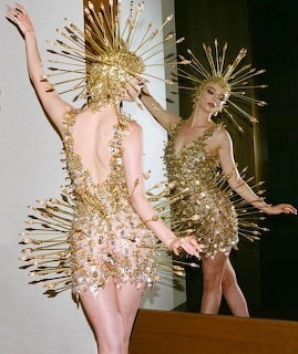 Woman in gold spiked dress and headpiece posing in front of a mirror