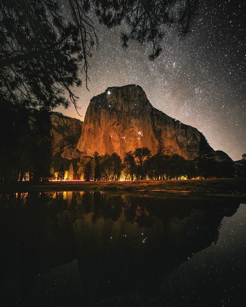 Large illuminated rock formation reflected in calm water under a starry night sky with glowing trees