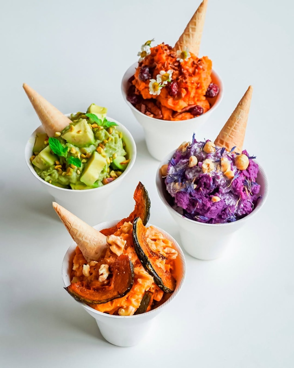 Four white cups with colorful food toppings and waffle cones arranged on a white surface
