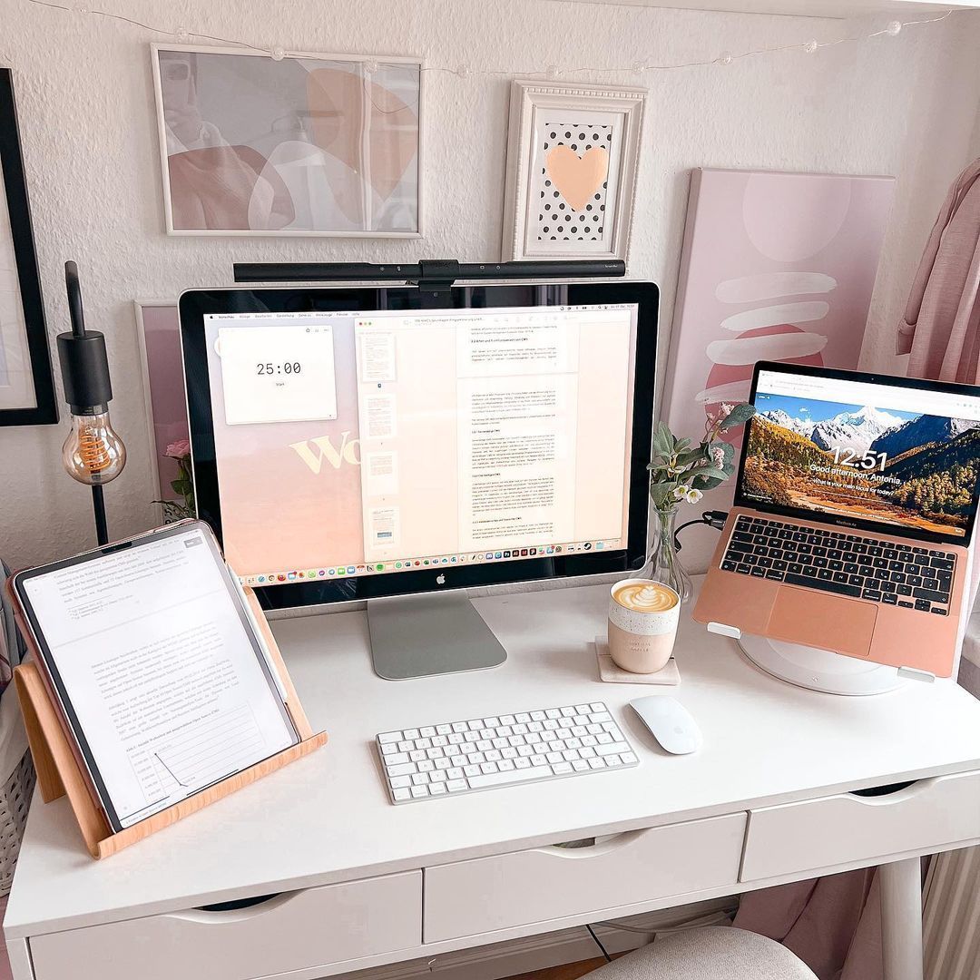 Modern minimalist home office desk setup with computer monitors, laptop, keyboard, and coffee mug in pink and white tones