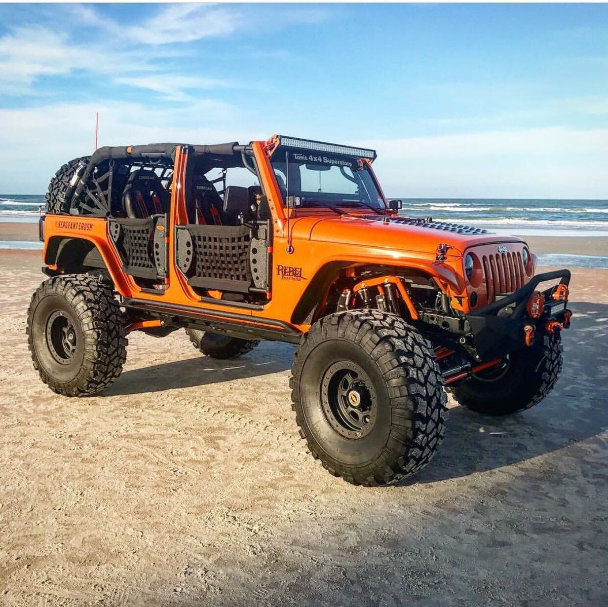 Orange Jeep Wrangler with oversized tires on beach sand near ocean