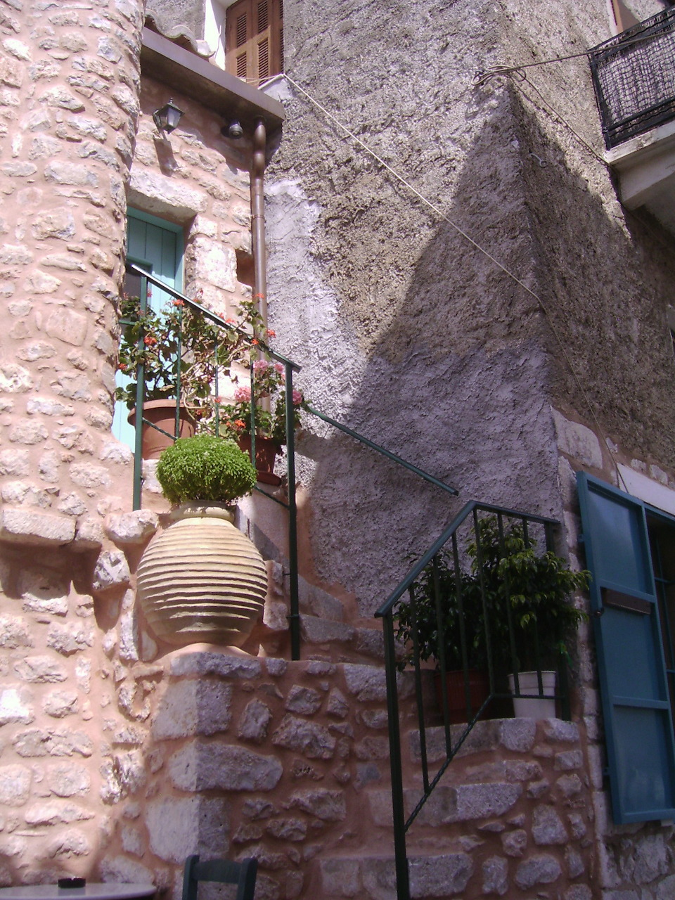 Sunlit narrow alley with stone walls, large terracotta urn, and potted plants in a Greek village
