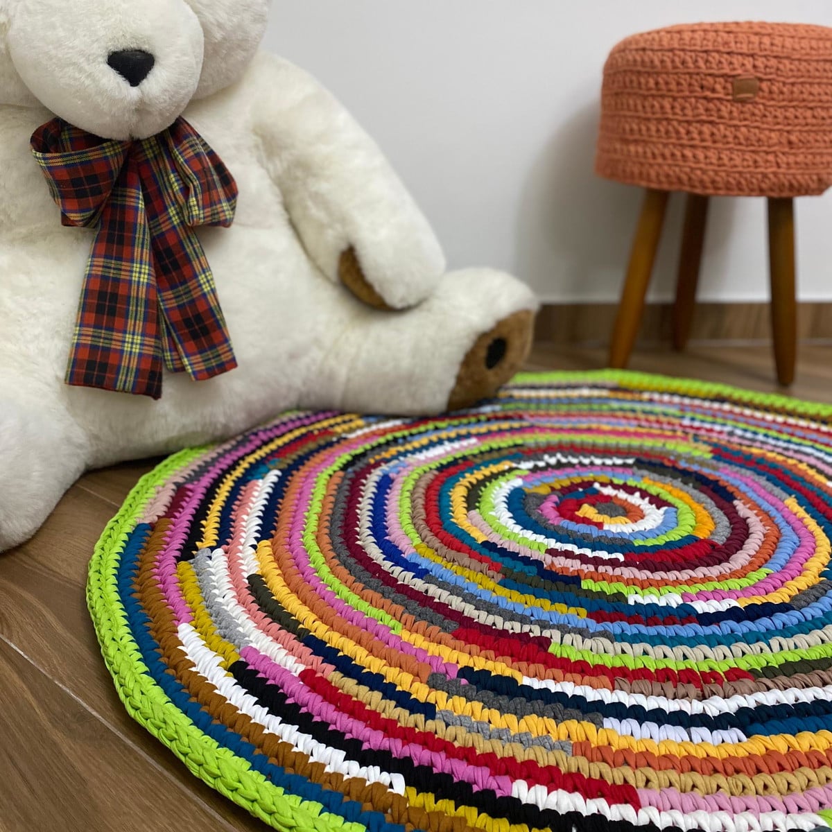 Colorful round crocheted rug on a wooden floor beside a white teddy bear and small stool