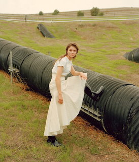 Woman in white dress standing by black hay bales in countryside field