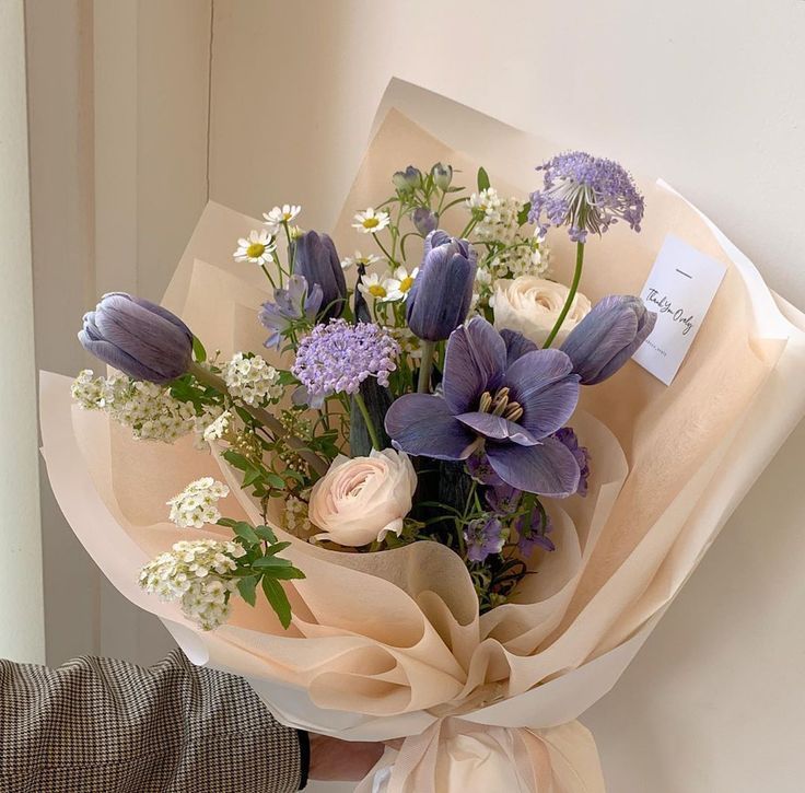 Hand holding wrapped bouquet with purple and cream flowers in translucent paper