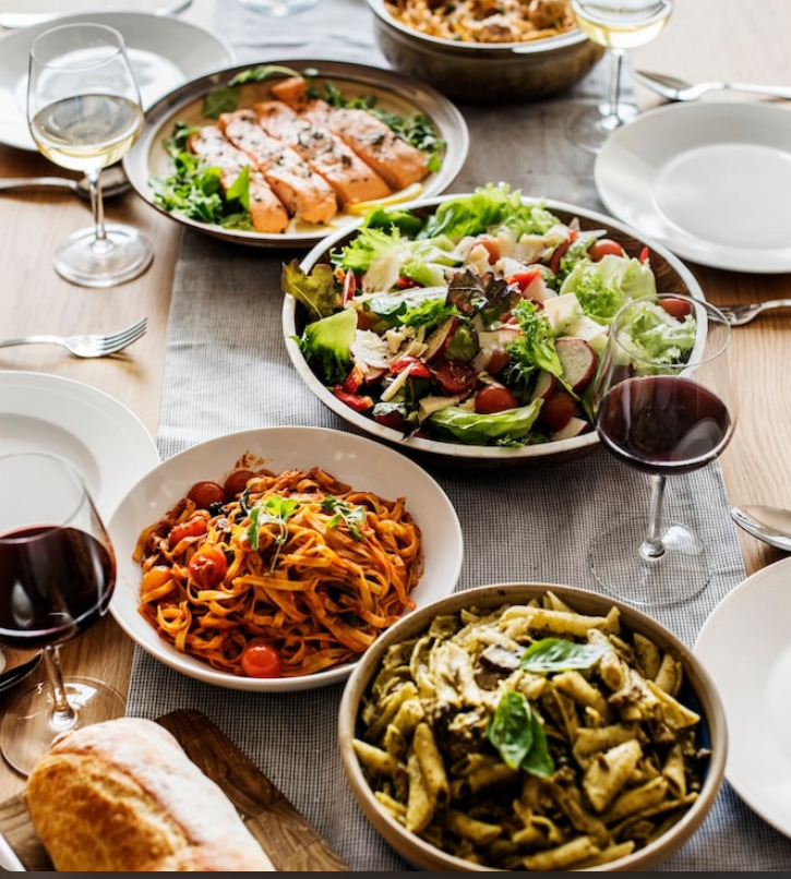 Dinner table with pasta dishes, salads, grilled chicken, and red wine glasses on a wooden surface