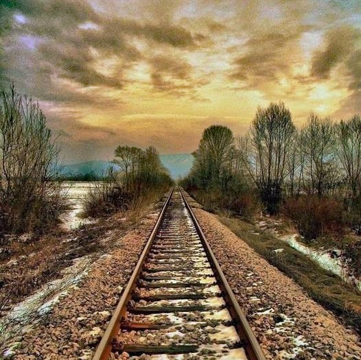 Railroad tracks leading toward sunset with dramatic cloudy sky and bare trees on both sides