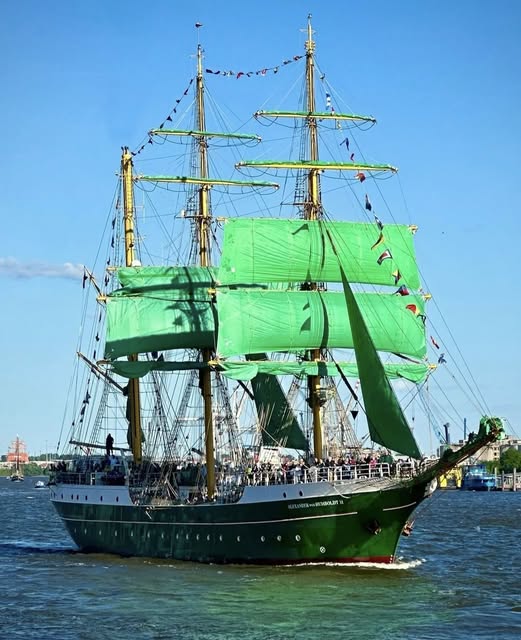 Green tall ship with full green sails sailing on open water near a city harbor