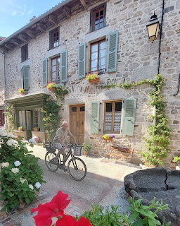 Stone building with green shutters and flower boxes, two bicycles parked in a coblestone courtyard