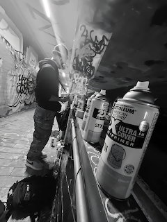Person spray painting graffiti in a dimly lit urban tunnel with spray cans lined up on a railing