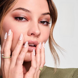 Close-up portrait of woman with white manicured nails touching face, wearing gold rings and subtle makeup