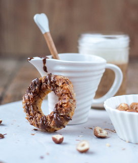 Nut-coated glazed pastry ring on a plate beside a white coffee cup and hazelnuts