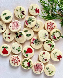 Round white chocolate cookies decorated with red flowers, green herbs, and berries arranged with fresh pine branches