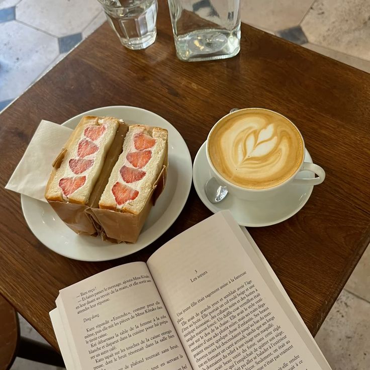 Latte art coffee, strawberry cream sandwich, and an open book on a wooden café table