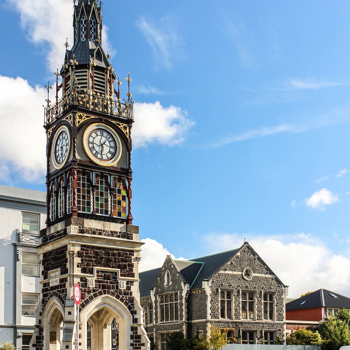 Victorian Gothic clock tower with ornate stonework and adjacent historic buildings under blue sky with white clouds