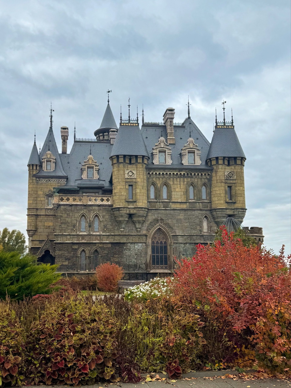 Medieval stone castle with multiple towers surrounded by red and orange autumn trees under cloudy sky