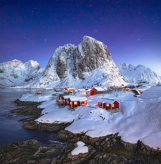 Snow-covered mountain peaks tower over traditional red cabins on a winter fjord under starlit sky