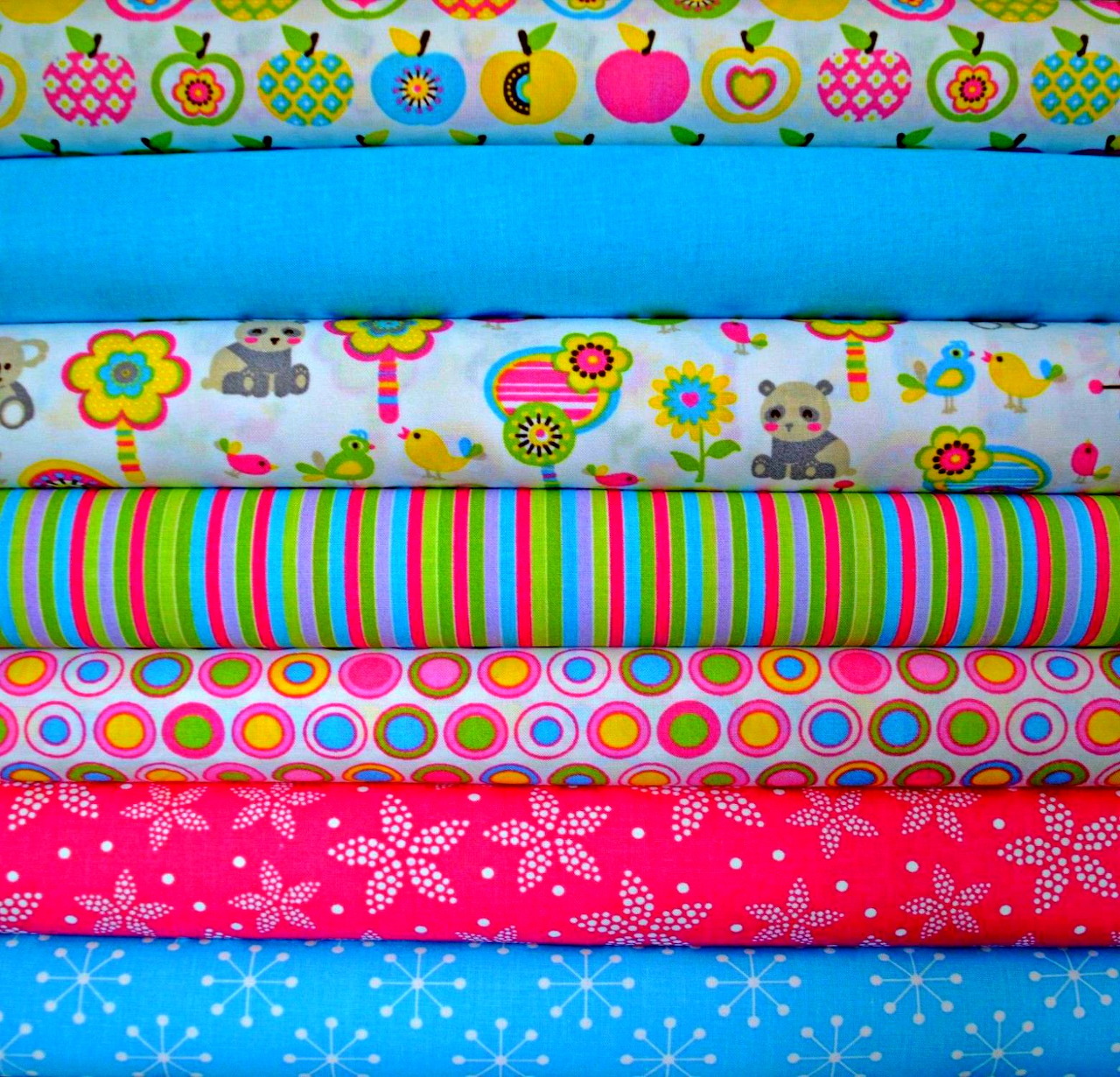 Six colorful patterned fabric rolls stacked horizontally showing apples, bears, flowers, stripes, circles and snowflakes