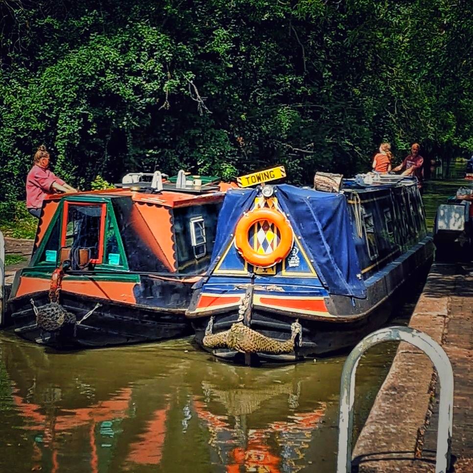 Two colorful narrowboats moored on a calm canal beside a tree-lined bank