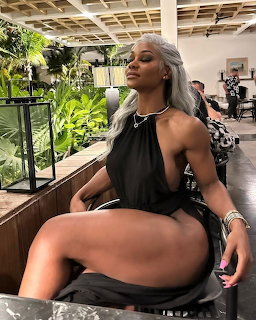 Muscular woman with silver hair in black halter dress posing seated in a salon with green plants behind her