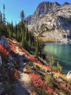 Hiking trail with red autumn shrubs beside a turquoise alpine lake with rocky mountain peaks and evergreen trees