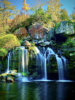 Multi-tiered waterfall flowing over mossy rocks into pool, framed by colorful fall foliage and arched stone bridge