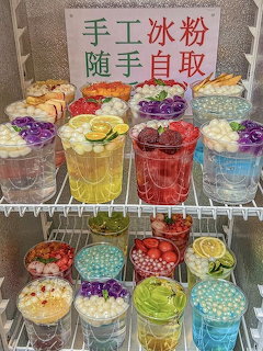Rows of colorful Chinese bing fen dessert drinks in clear cups with fruit and topping garnishes on refrigerator shelves.