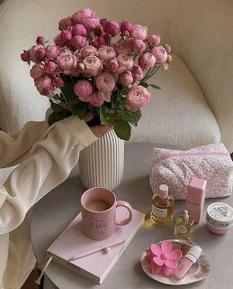 Pink peony bouquet in white vase with pink mug, books, and beauty products on a table