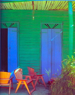 Green wooden house with blue doors, yellow awning, and red chairs on patio
